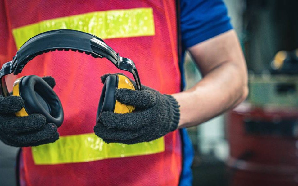 close up of man holding a pair of noise protection earmuffs wearing a safety best and protective gloves inside an industrial environment