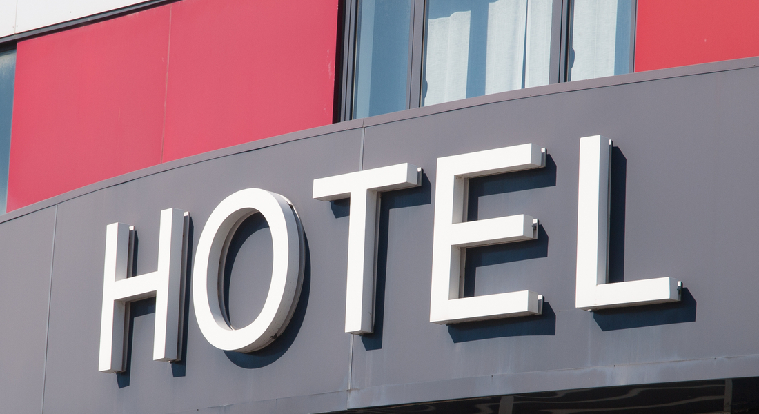 The façade of a hotel building has several windows lining the front between red walls, and a large sign reads "HOTEL."