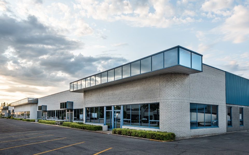 The exterior of a brick commercial building with large windows, a big parking lot, and shrubs and bushes by the front entrance.