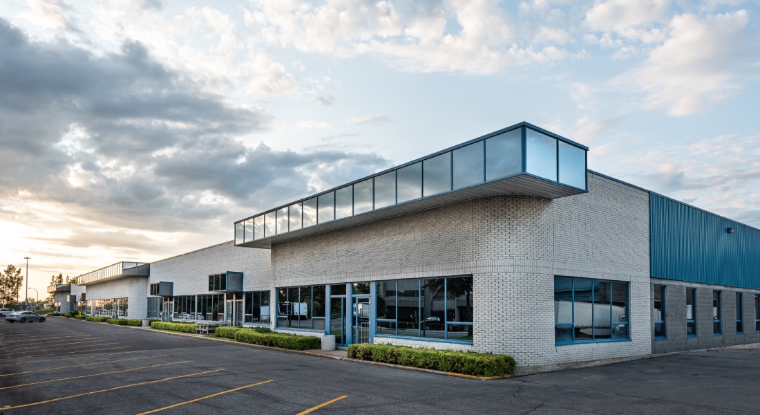 The exterior of a brick commercial building with large windows, a big parking lot, and shrubs and bushes by the front entrance.