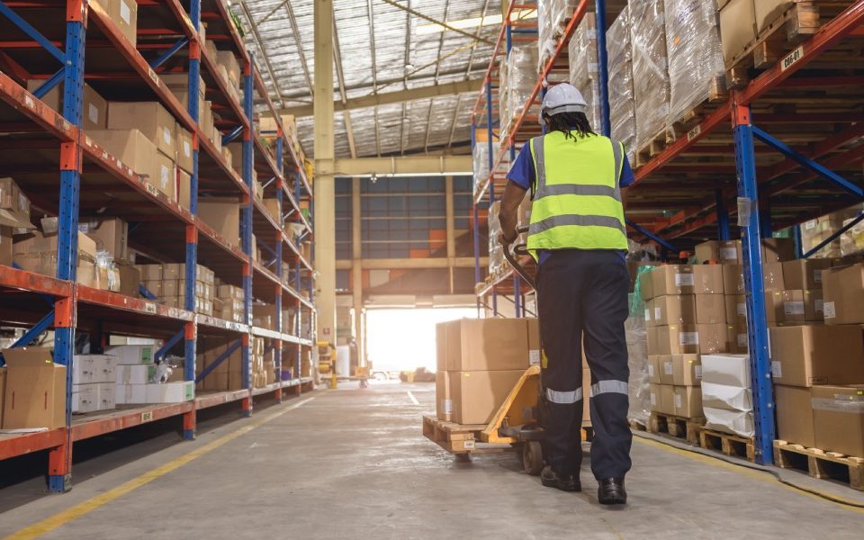 Worker wearing a safety vest pushing a pallet with multiple boxes down a large aisle inside a shipping warehouse.