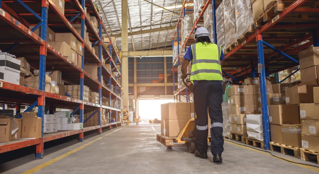 Worker wearing a safety vest pushing a pallet with multiple boxes down a large aisle inside a shipping warehouse.