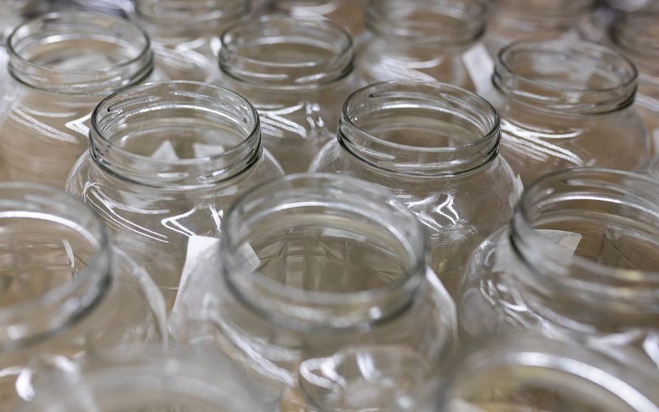 A bulk supply of lidless glass jars lines a shelf. They are made of clear glass and have unreadable white labels.