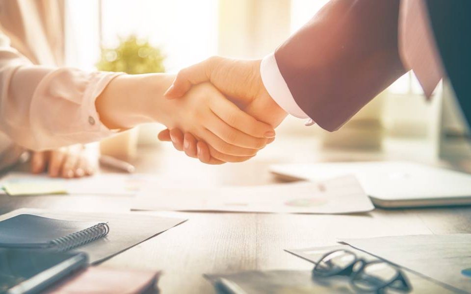 A man and a woman shaking hands in a glowing, sunlit office. The table between them holds various office essentials.