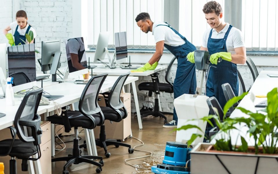 A male and a female are cleaning a commercial office space. One person is waxing the floors, and the others are wiping counters.