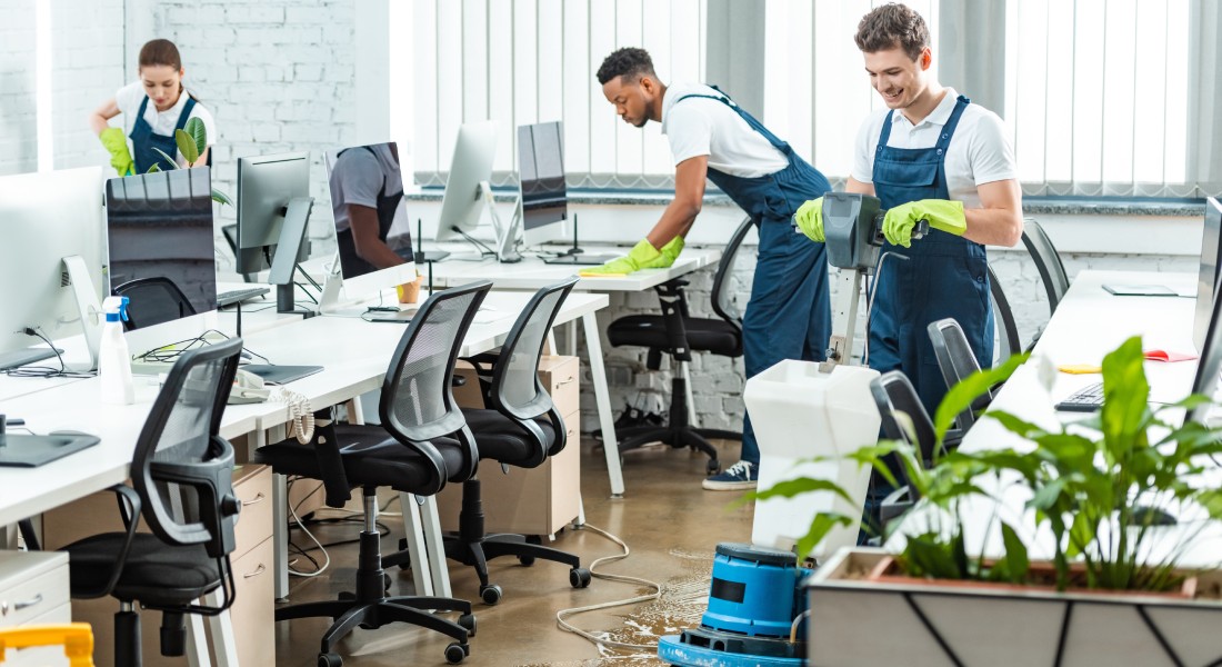 A male and a female are cleaning a commercial office space. One person is waxing the floors, and the others are wiping counters.