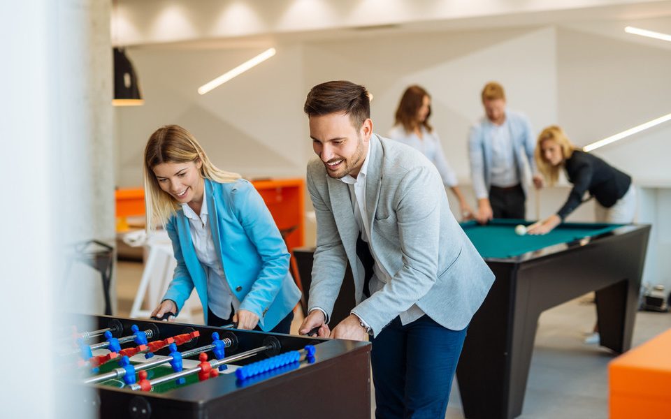 A man and a woman in semiformal clothing playing foosball together as a group of three plays pool behind them.