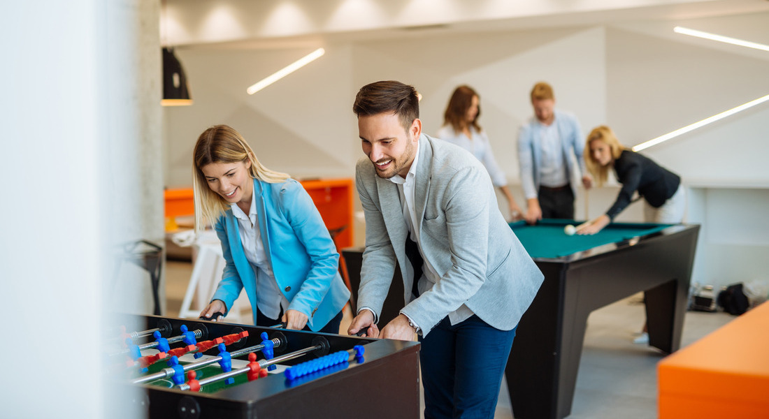 A man and a woman in semiformal clothing playing foosball together as a group of three plays pool behind them.