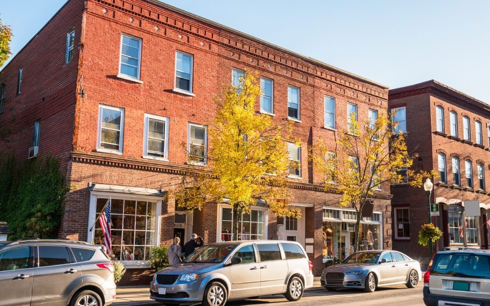 The exterior of a multi-level brick building with storefronts on the street level. There are cars parked along the street.