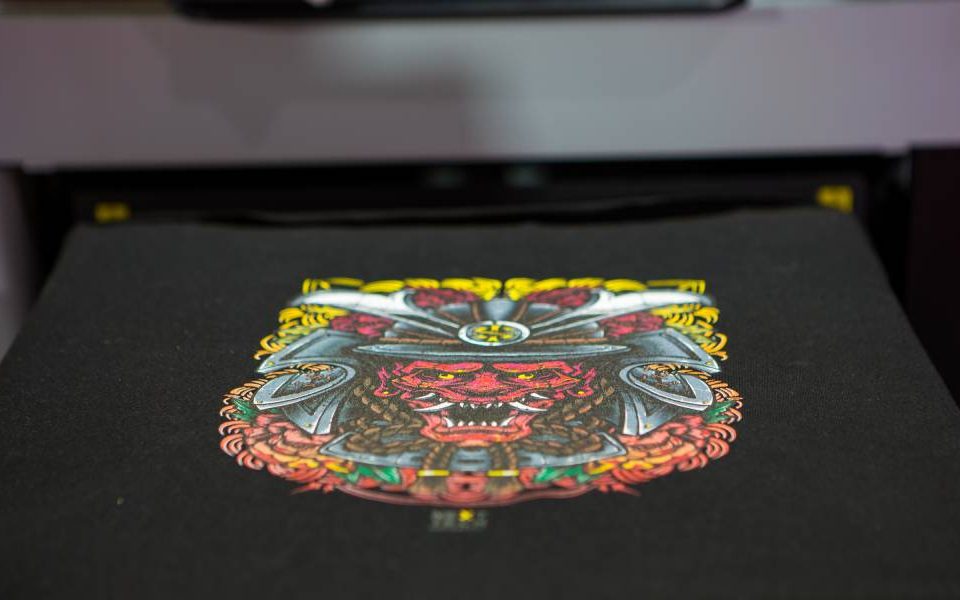 A garment printer is printing out a vibrant, detailed graphic of a red hannya mask onto a black t-shirt.