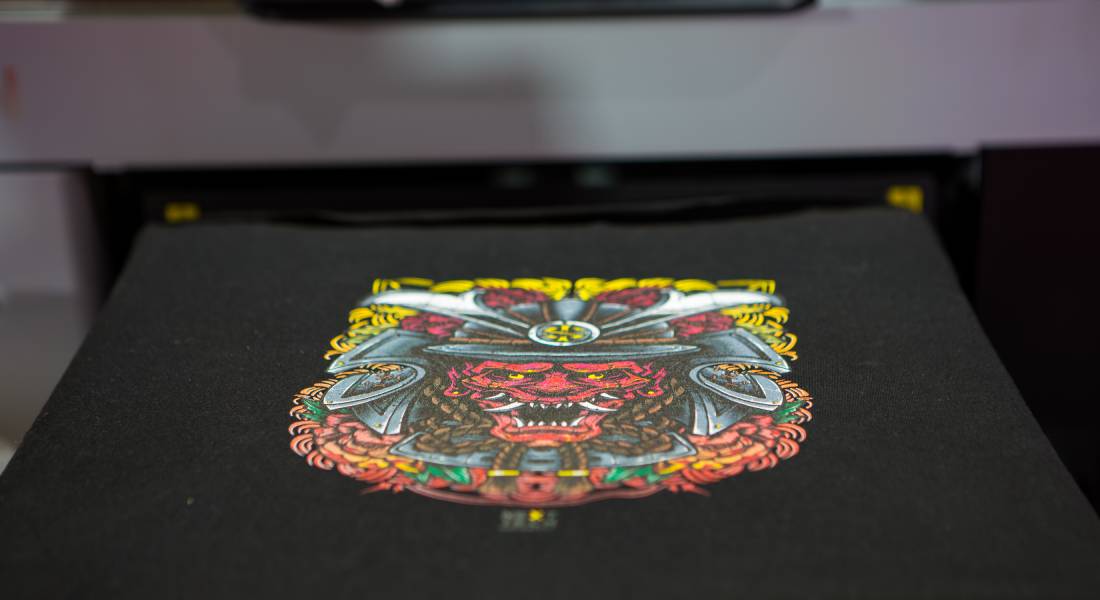 A garment printer is printing out a vibrant, detailed graphic of a red hannya mask onto a black t-shirt.