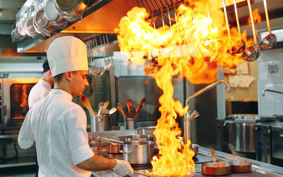 A chef in a commercial kitchen is wearing a white hat and gloves while holding a flaming pan by the handle.