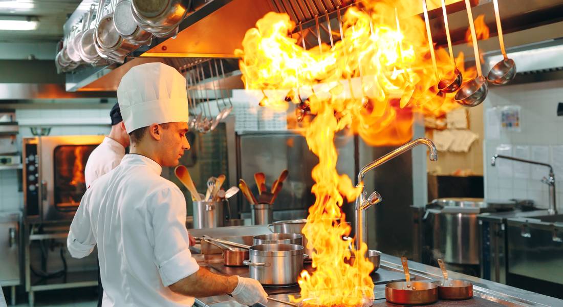 A chef in a commercial kitchen is wearing a white hat and gloves while holding a flaming pan by the handle.