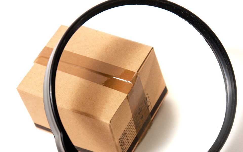 A hand holding a magnifying glass over a miniature cardboard shipping box sealed with packing tape.