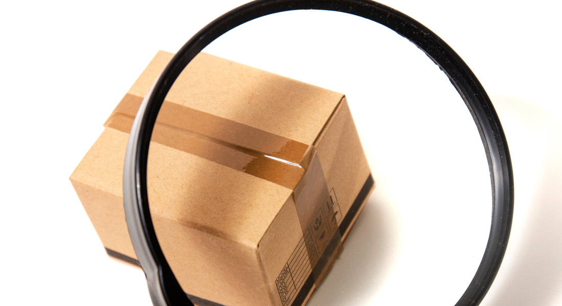 A hand holding a magnifying glass over a miniature cardboard shipping box sealed with packing tape.