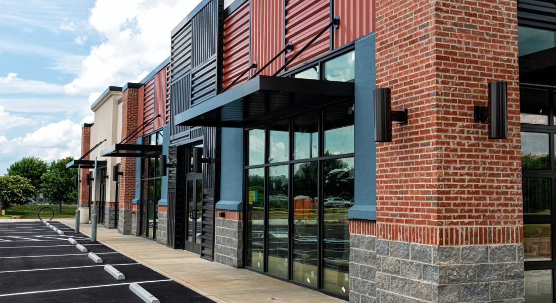 A business building contains a black metal, red brick, and tan stone exterior. The parking spaces are empty.