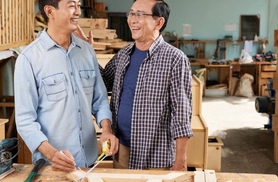 Two men standing beside a woodworking table. One holds an extended tape measure in one hand and a pencil in the other.