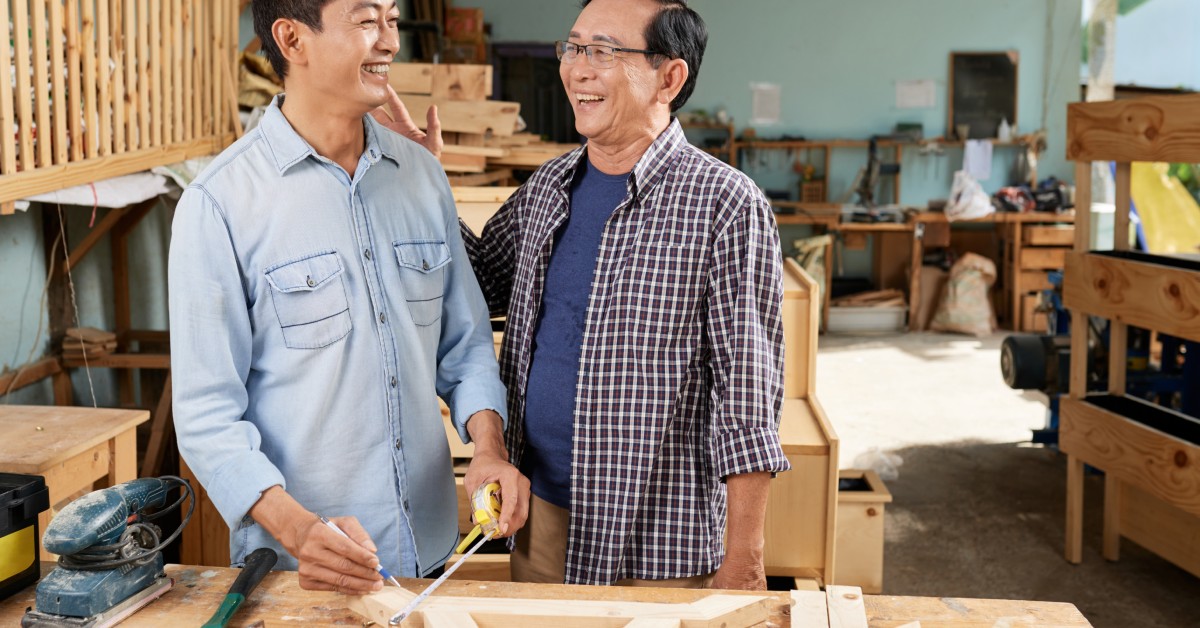 Two men standing beside a woodworking table. One holds an extended tape measure in one hand and a pencil in the other.