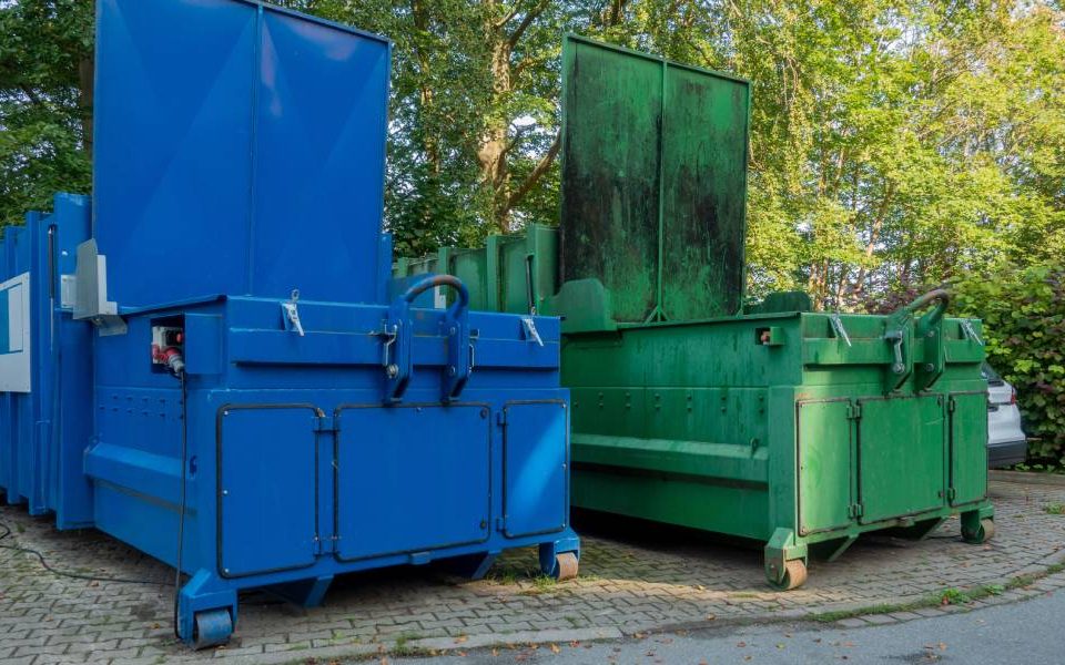 Two large waste compactors next to each other with open loading doors placed in an outdoor environment with trees in background.