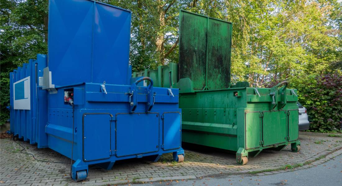 Two large waste compactors next to each other with open loading doors placed in an outdoor environment with trees in background.