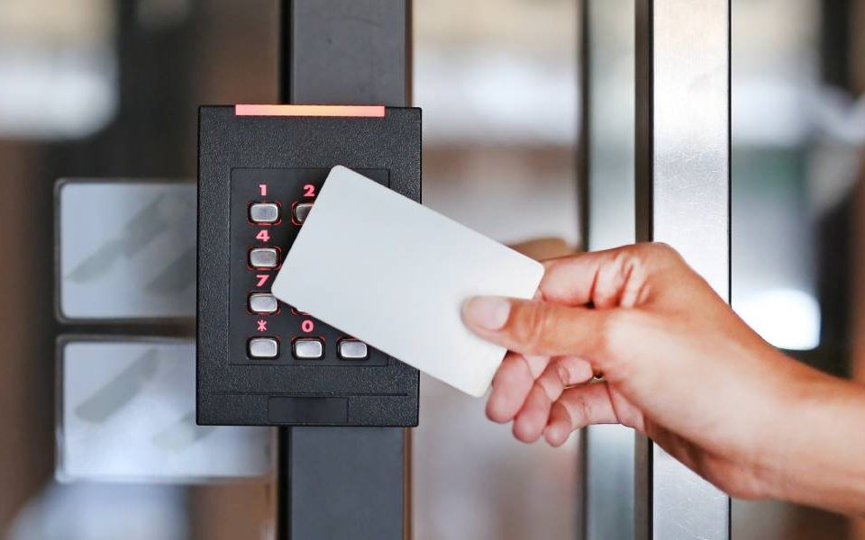 A hand is holding a white keycard up to the access control system on the door of a building to unlock it.