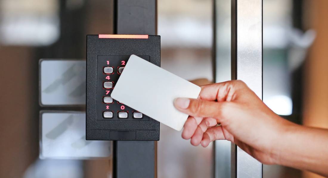 A hand is holding a white keycard up to the access control system on the door of a building to unlock it.