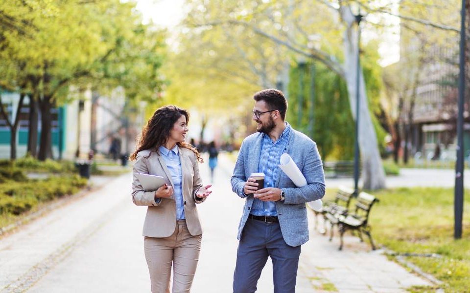 A man and woman walk together outdoors in a public space. They are wearing business attire and face each other in conversation.