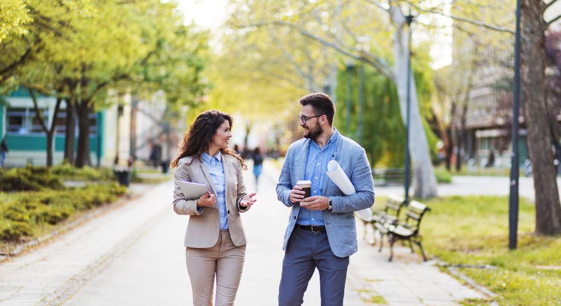 A man and woman walk together outdoors in a public space. They are wearing business attire and face each other in conversation.