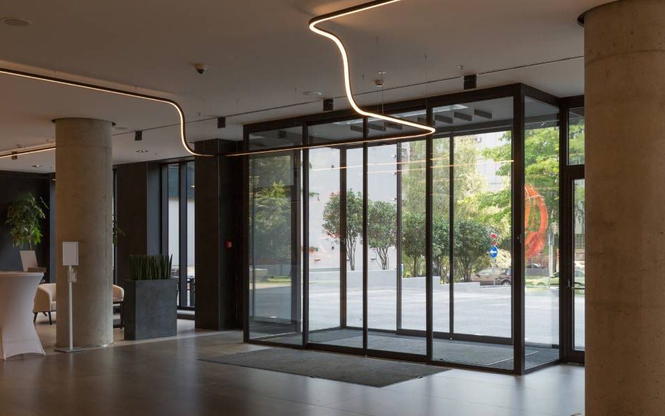 The lobby of a modern building with concrete columns and accent lighting. The entryway has automatic glass sliding doors.