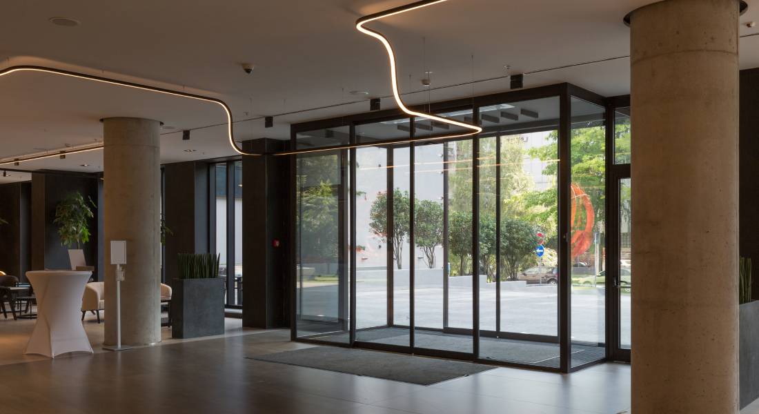 The lobby of a modern building with concrete columns and accent lighting. The entryway has automatic glass sliding doors.