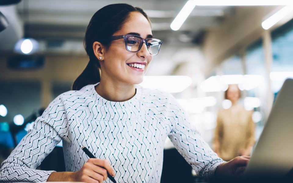 Woman wearing glasses and a white top sitting behind a desk holding a pen and smiling while looking at a laptop screen.