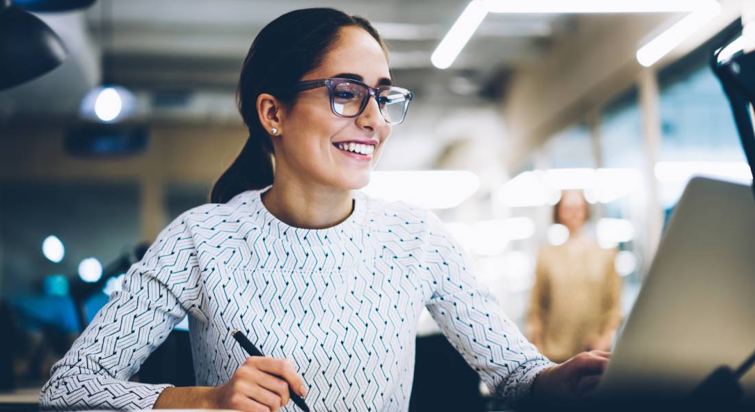 Woman wearing glasses and a white top sitting behind a desk holding a pen and smiling while looking at a laptop screen.