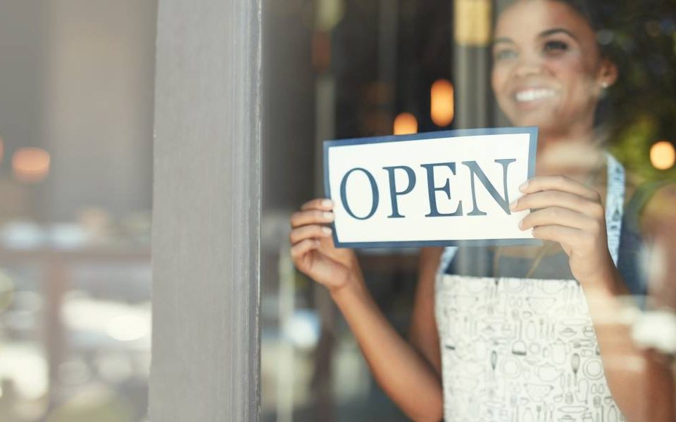 A woman in a white apron with designs on it is smiling through a window and setting up an "OPEN" sign.