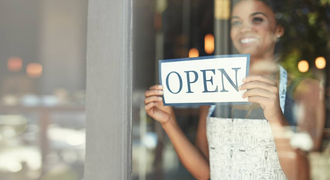 A woman in a white apron with designs on it is smiling through a window and setting up an "OPEN" sign.