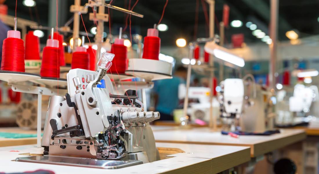 An overlock sewing machine with multiple cones of red thread and a straight sewing machine sit inside a warehouse.